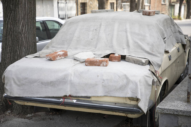 Старый автомобиль покрыт серым брезентом, кирпичами и камнями
 - Фото, изображение
