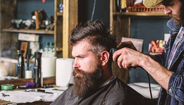 Hipster client getting haircut. Haircut concept. Barber styling hair of bearded client with comb and clipper. Barber with hair clipper works on hairstyle for man with beard, barbershop background - Foto, Imagen