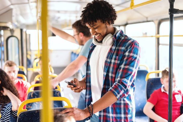 若い笑顔アフリカ系アメリカ人の男スマートフォンを使用して、市内バスに乗りながら立って. - 写真・画像