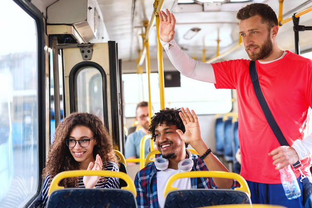 Felices amigos multiculturales saludando a alguien mientras está sentado y de pie en el autobús de la ciudad
. - Foto, imagen