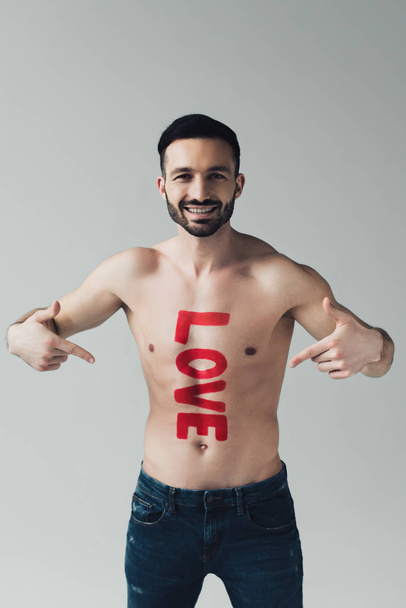 front view of smiling shirtless man pointing with fingers at inscription on body isolated on grey - Foto, Imagen
