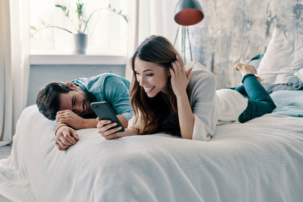 Piękna młoda para korzystająca ze smartfona i uśmiechnięta spędzając czas w łóżku w domu    - Zdjęcie, obraz