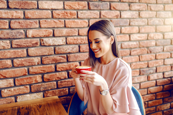 Retrato de una joven caucásica disfrutando de delicioso café cremoso mientras está sentada en la cafetería. En la pared de ladrillo de fondo
. - Foto, Imagen