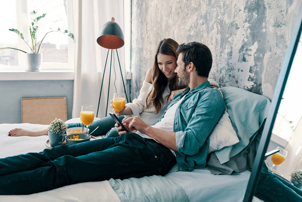 Caucasian couple having breakfast while spending time in bed at home. browsing mobile phone and having breakfast with orange juice  - Zdjęcie, obraz
