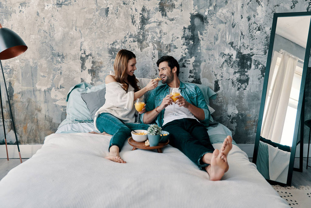 Caucasian couple having breakfast with orange juice while spending time in bed at home, woman feeding man              - Foto, Bild