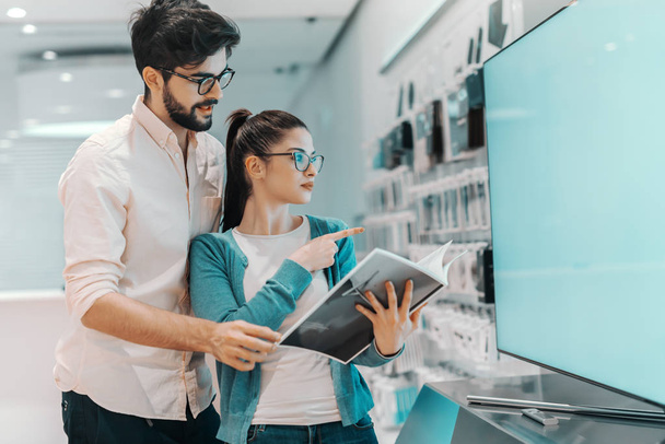 Young attractive multicultural couple looking for new plasma tv. Woman holding brochure and pointing at tv. - Photo, Image