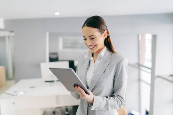 Smiling Caucasian female CEO in formal wear and with long brown hair using tablet while standing at office. Small opportunities are often the beginning of great achievements. - Фото, зображення