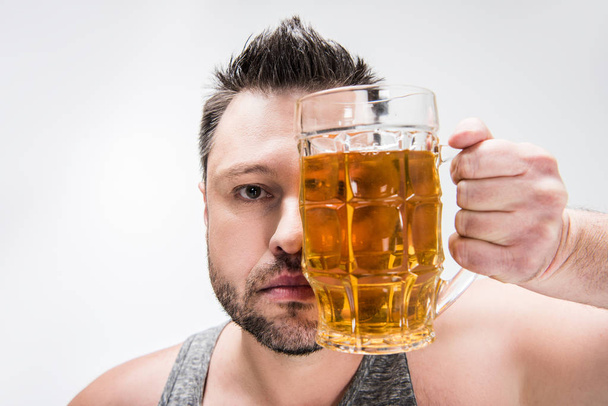 υπέρβαρο άνθρωπος κοιτάζοντας την κάμερα και καλύπτοντας το πρόσωπο με ποτήρι μπύρας απομονωθεί σε λευκό - Φωτογραφία, εικόνα