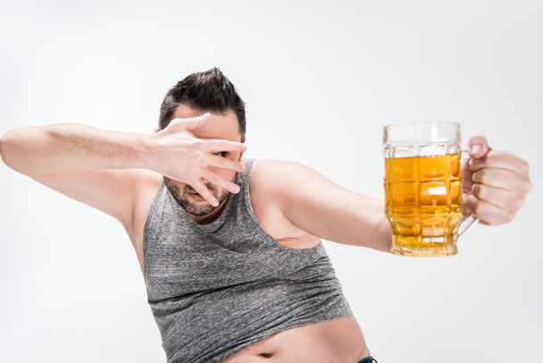 overweight man covering face with hand and holding glass of beer isolated on white - Фото, изображение