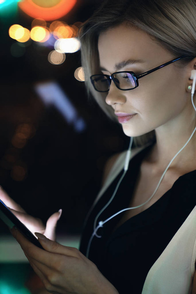 Vertical shot of young beautiful woman sending email with tablet in her hand and earphones lale night in office with lights on background - Fotoğraf, Görsel