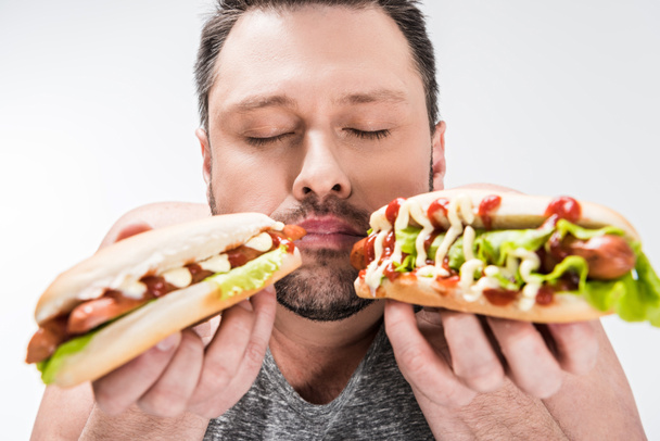 übergewichtiger Mann mit geschlossenen Augen, der leckere Hot Dogs auf weißem Eis hält - Foto, Bild