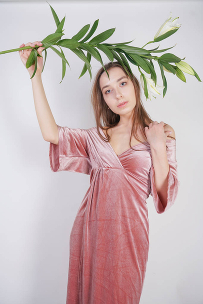 shy caucasian girl with big eyes stands in a velvet pink dress and holds a white Lily in her hand as a symbol of innocence and purity. young woman posing on white studio background. - Φωτογραφία, εικόνα