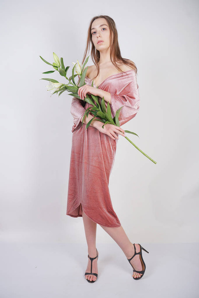 shy caucasian girl with big eyes stands in a velvet pink dress and holds a white Lily in her hand as a symbol of innocence and purity. young woman posing on white studio background. - Photo, Image
