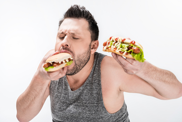 éhes túlsúlyos ember a tank Top evés hot dog izolált fehér - Fotó, kép