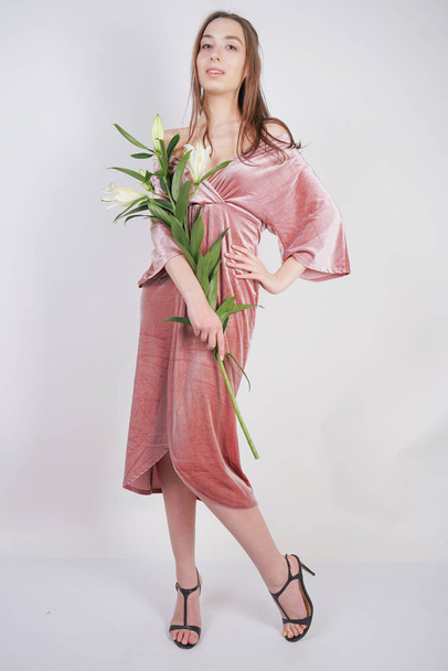 shy caucasian girl with big eyes stands in a velvet pink dress and holds a white Lily in her hand as a symbol of innocence and purity. young woman posing on white studio background. - Foto, immagini