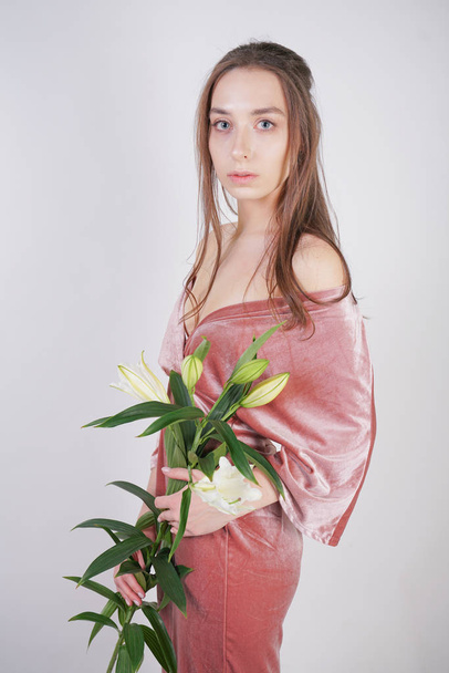 shy caucasian girl with big eyes stands in a velvet pink dress and holds a white Lily in her hand as a symbol of innocence and purity. young woman posing on white studio background. - Photo, Image