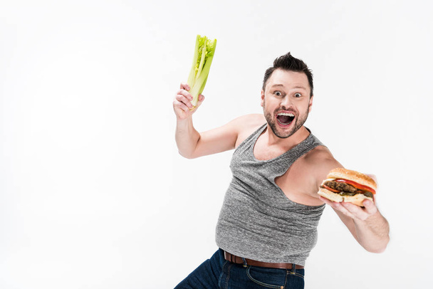 ενθουσιασμένος υπέρβαρων άνθρωπος κρατώντας σέλινο και burger απομονωθεί σε λευκό με αντίγραφο χώρου - Φωτογραφία, εικόνα