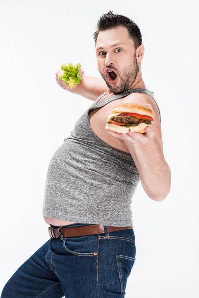 збуджений чоловік з надмірною вагою дивиться на камеру, тримаючи селеру та бургер ізольовано на білому
  - Фото, зображення