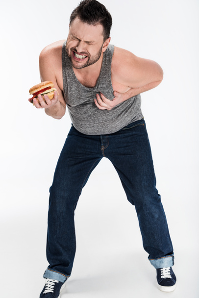 υπέρβαρο άνθρωπος στην κορυφή της δεξαμενής κρατώντας burger και αγγίζοντας το στήθος σε λευκό - Φωτογραφία, εικόνα
