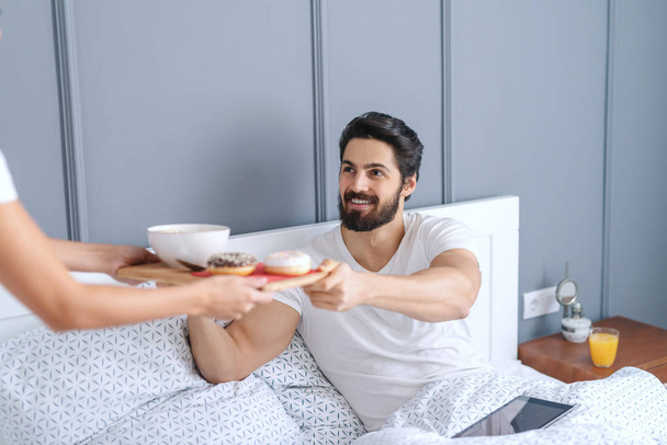 Жінка приносить сніданок своєму люблячому чоловікові. Чоловік сидить у ліжку і бере лоток
. - Фото, зображення