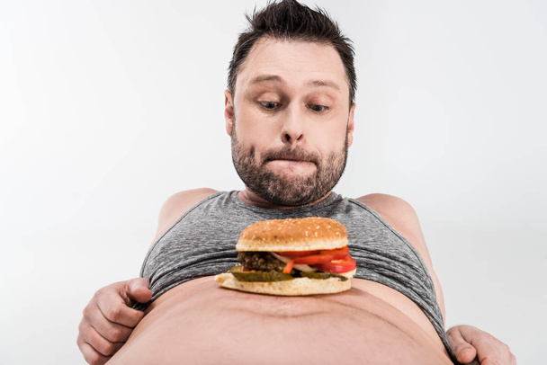 hombre con sobrepeso mirando hamburguesa en el vientre aislado en blanco
 - Foto, imagen
