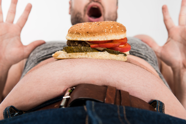 částečný pohled na vzrušeného nadváhy muže s hamburgerem na břichu gestikuloval izolovaný na bílém - Fotografie, Obrázek