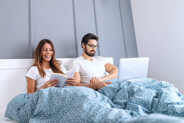 Smiling beautiful Caucasian brunette reading book in bed while her husband with eyeglasses using laptop. - Photo, Image