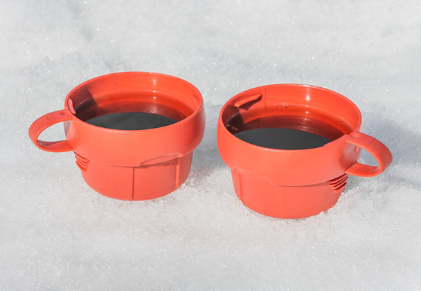 Две красные пластиковые кружки с черным горячим чаем с паром на белом фоне снега на улице зимой для пикника
 - Фото, изображение
