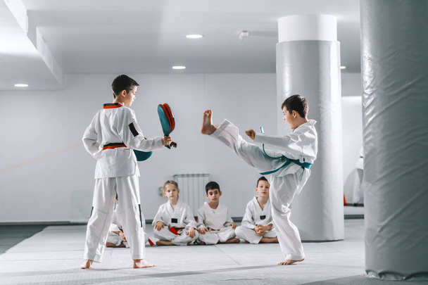 Δύο νεαρά Καυκάσια αγόρια σε doboks έχουν taekwondo εκπαίδευση στο γυμναστήριο. Το ένα αγόρι κλωτσάει ενώ το άλλο κρατάει το στόχο. Στο παρασκήνιο ο φίλος τους κάθεται με τα πόδια σταυρωμένα και βλέποντας τους. - Φωτογραφία, εικόνα