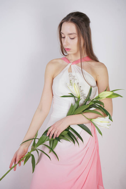 a charming young girl with big eyes and brown hair stands without makeup in a fashion dress on a white background in the Studio. beautiful young teen woman holding a stem with white lush lilies. - Фото, изображение