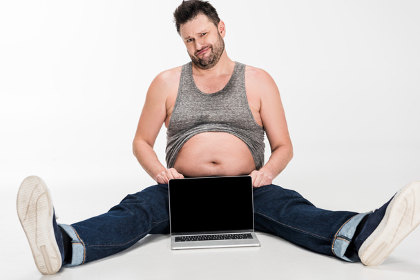 skeptical overweight man looking at camera while sitting near laptop with blank screen on white - Foto, Bild