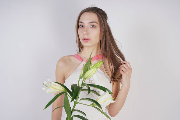 Ein charmantes junges Mädchen mit großen Augen und braunen Haaren steht ungeschminkt in einem Modekleid vor weißem Hintergrund im Atelier. schöne junge Teen-Frau hält einen Stiel mit weißen üppigen Lilien. - Foto, Bild