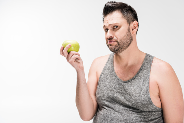 niezadowolony z nadwagą człowiek trzyma zielone jabłko i patrząc na aparat na białym tle - Zdjęcie, obraz