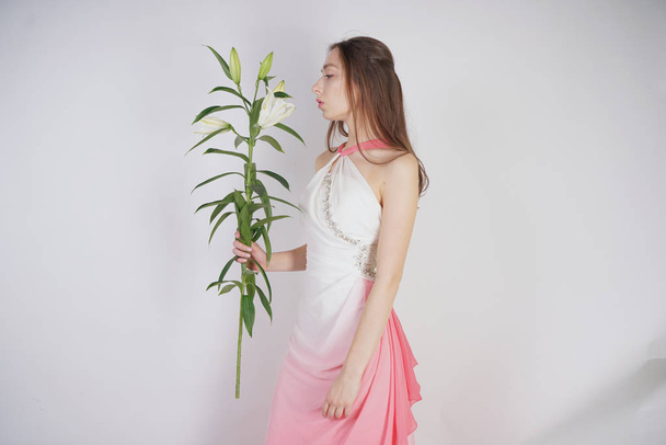a charming young girl with big eyes and brown hair stands without makeup in a fashion dress on a white background in the Studio. beautiful young teen woman holding a stem with white lush lilies. - Photo, Image