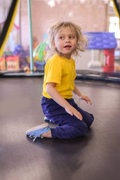 Retrato de un chico rubio con una camiseta amarilla. El niño sonríe y juega en la sala de juegos de los niños. El niño salta en el trampolín
. - Foto, Imagen
