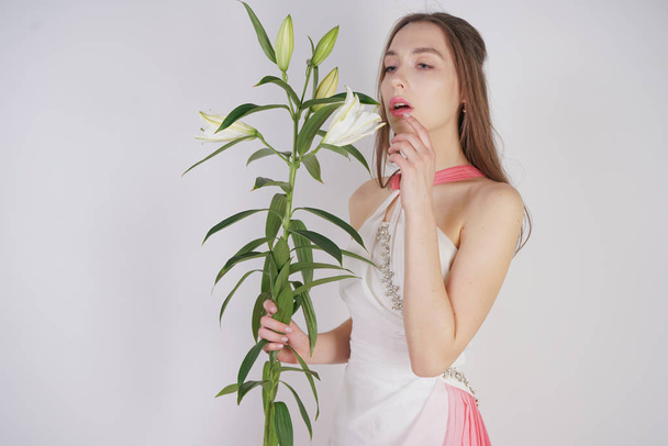 a charming young girl with big eyes and brown hair stands without makeup in a fashion dress on a white background in the Studio. beautiful young teen woman holding a stem with white lush lilies. - Fotó, kép