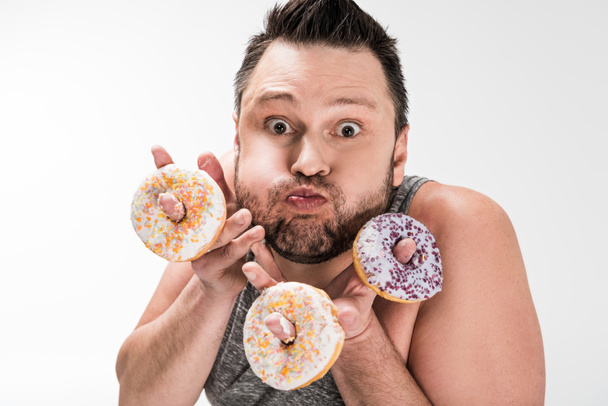 gordito hombre haciendo expresión de la cara mientras sostiene donuts y mirando a la cámara aislado en blanco
 - Foto, imagen