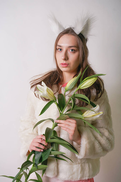 cute young teen girl standing in a winter warm fur coat and cat furry ears on her head, she holds a branch of fresh lilies. woman in winter clothes with a flower on a white background in the Studio. - Photo, Image