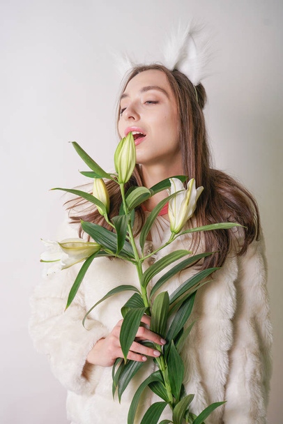 cute young teen girl standing in a winter warm fur coat and cat furry ears on her head, she holds a branch of fresh lilies. woman in winter clothes with a flower on a white background in the Studio. - Fotoğraf, Görsel