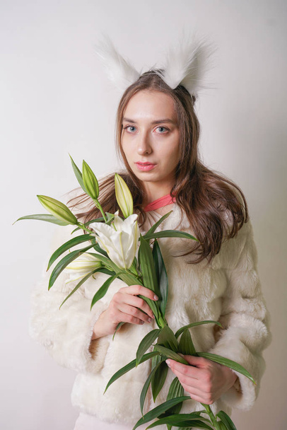 彼女の頭の上の冬暖かい毛皮のコートと猫の毛皮の耳に立ってかわいい若い十代の少女は、彼女は新鮮なユリの枝を保持しています。スタジオの白い背景に花を咲かせる冬服の女. - 写真・画像