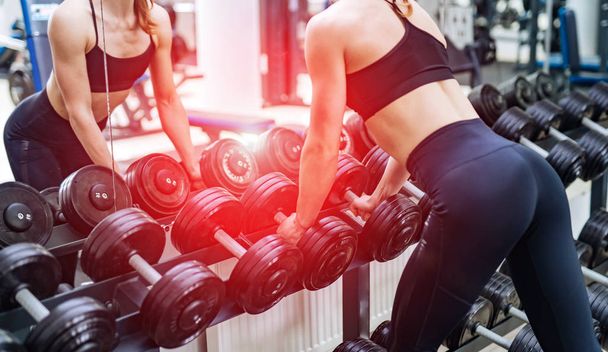 Muscular body of a girl leaning on the heavy dumbbells in front of the mirror in a sports hall. Strong young beautiful woman is taking sports equipment near the mirror. - Foto, immagini