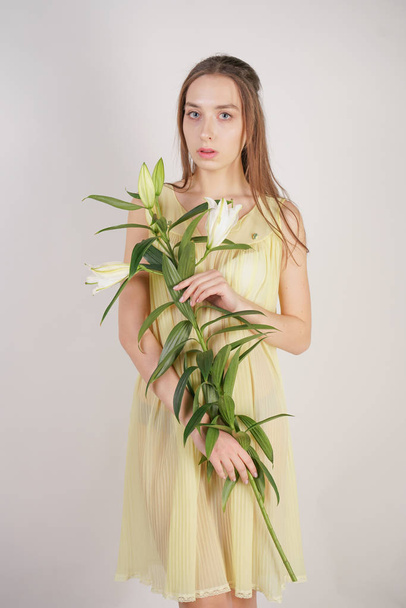 uma encantadora jovem caucasiana em um amarelo retro plissado nightie transparente segura uma exuberante flor Lily em suas mãos e fica em um fundo branco no estúdio
 - Foto, Imagem