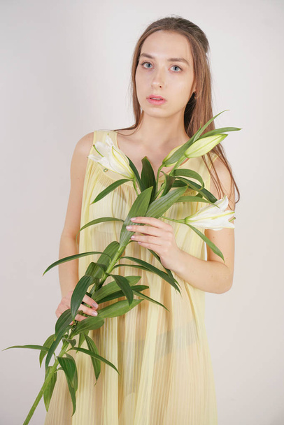 a charming young caucasian girl in a retro yellow pleated transparent nightie holds a lush Lily flower in her hands and stands on a white background in the Studio - Foto, Imagen