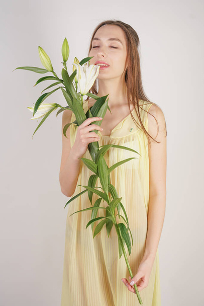 a charming young caucasian girl in a retro yellow pleated transparent nightie holds a lush Lily flower in her hands and stands on a white background in the Studio - Photo, Image