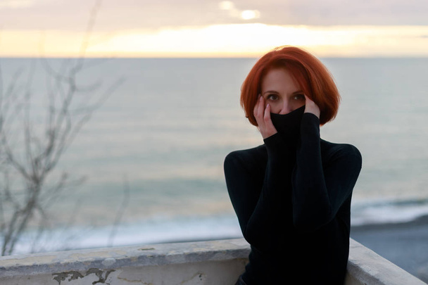 Portrét mladé ženy s červenými vlasy s neklidným pohledem na pozadí moře a západu slunce v jarním večeru - Fotografie, Obrázek