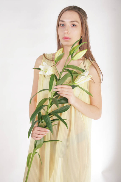 レトロな黄色のプリーツ透明のネグリジェで、美しい若い白人の女の子が彼女の手に緑豊かなユリの花を保持し、スタジオで白い背景の上に立っている - 写真・画像