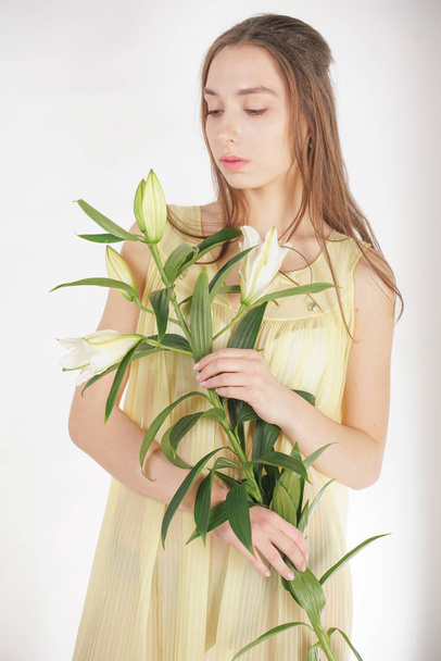 ένα γοητευτικό νεαρό καυκάσιο κορίτσι σε ένα ρετρό κίτρινο πλισέ διαφανές νυχτικό κρατά ένα πλούσιο λουλούδι Lily στα χέρια της και στέκεται σε ένα λευκό φόντο στο στούντιο - Φωτογραφία, εικόνα