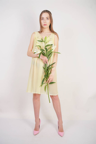 a charming young caucasian girl in a retro yellow pleated transparent nightie holds a lush Lily flower in her hands and stands on a white background in the Studio - Photo, image