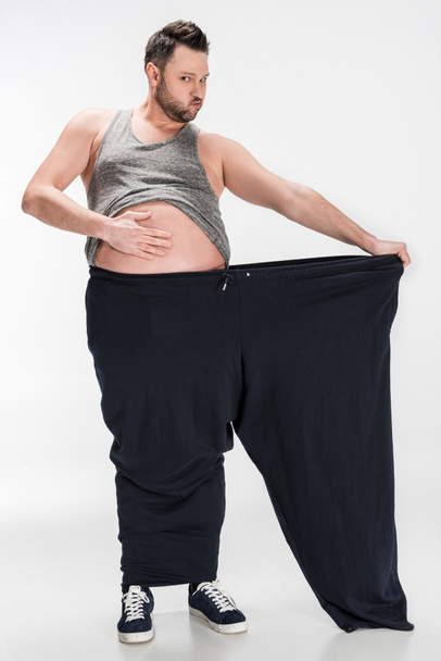 overweight man touching belly while holding oversize pants after weight loss on white - Foto, Imagem