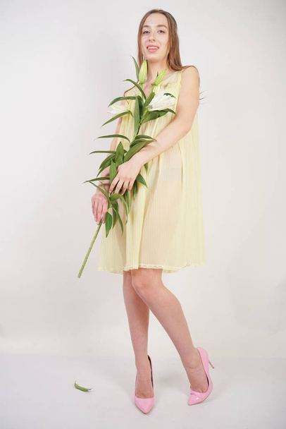 a charming young caucasian girl in a retro yellow pleated transparent nightie holds a lush Lily flower in her hands and stands on a white background in the Studio - Foto, afbeelding